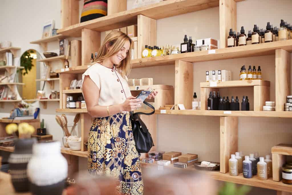 Female Customer Shopping In Independent Cosmetics Store Comparing Products Using Mobile Phone