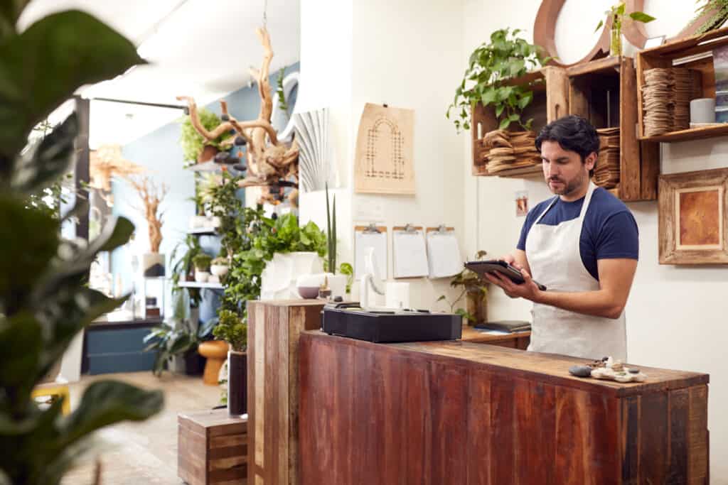 Male Owner With Digital Tablet Standing Behind Sales Desk Of Florists Store